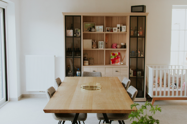 Moderne eetruimte met houten eettafel, zes stoelen en een open wandkast, met zicht op kinderbedje en veel daglicht.